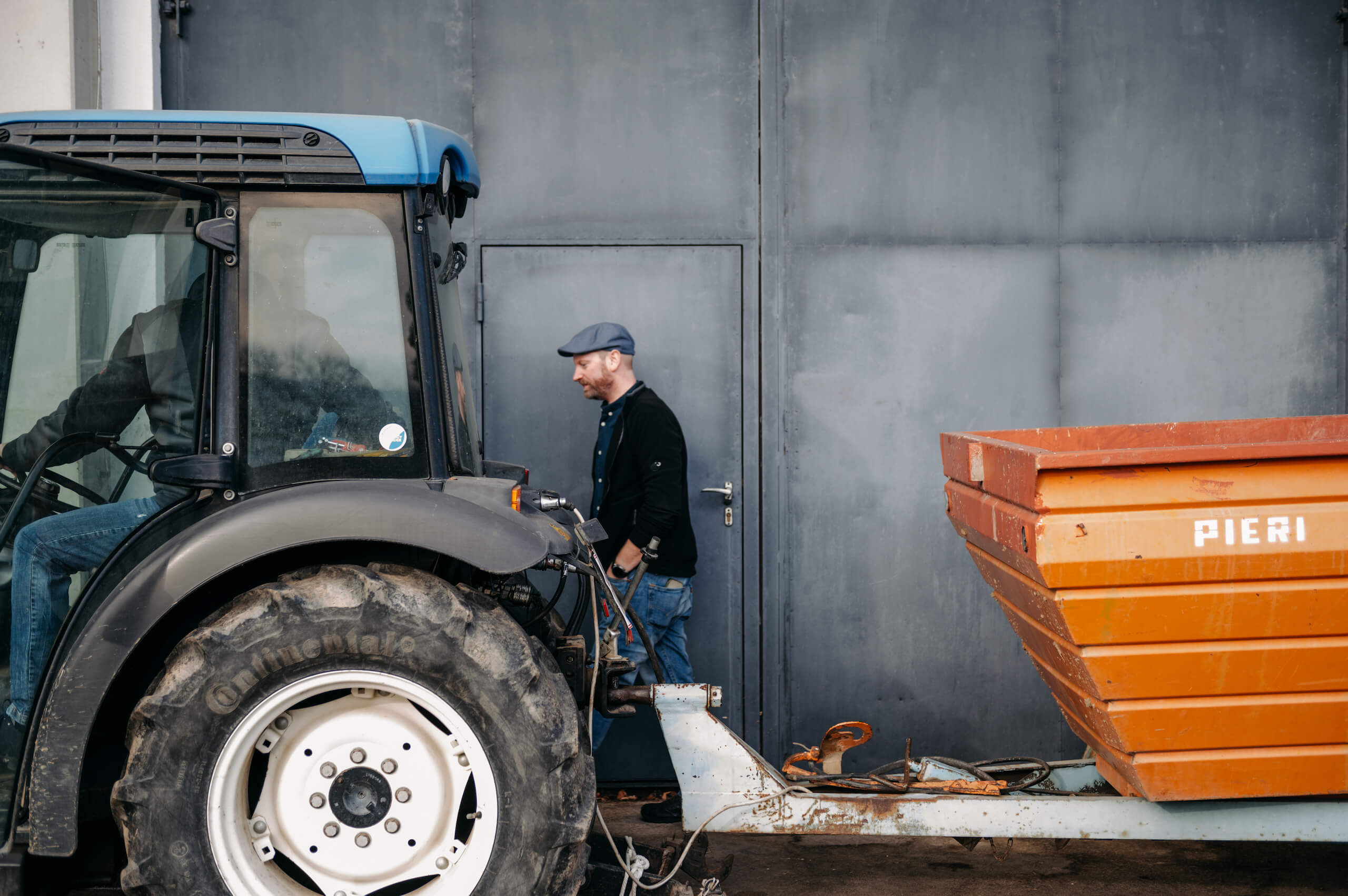 Thomas sitzt auf einem blauen Traktor mit einem orangen Anhänger und Alexander geht daneben vorbei