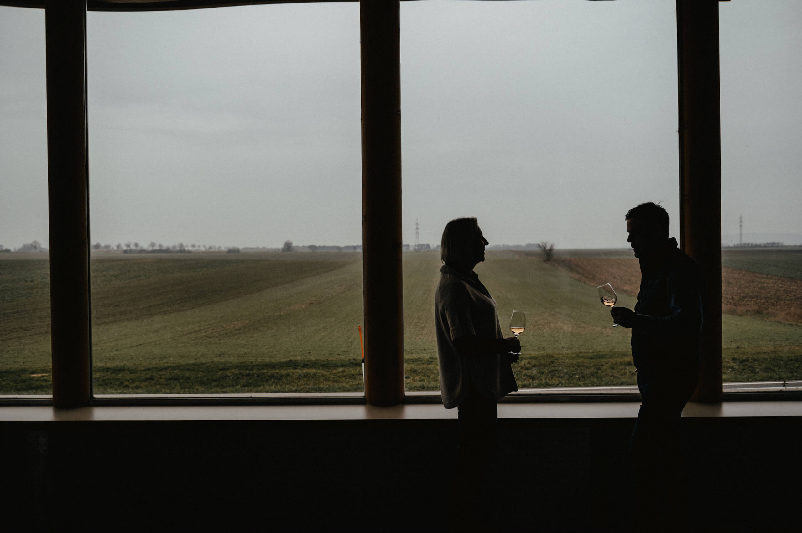 Erich und Anna Maria stehen vor einem großen Fenster und trinken Wein