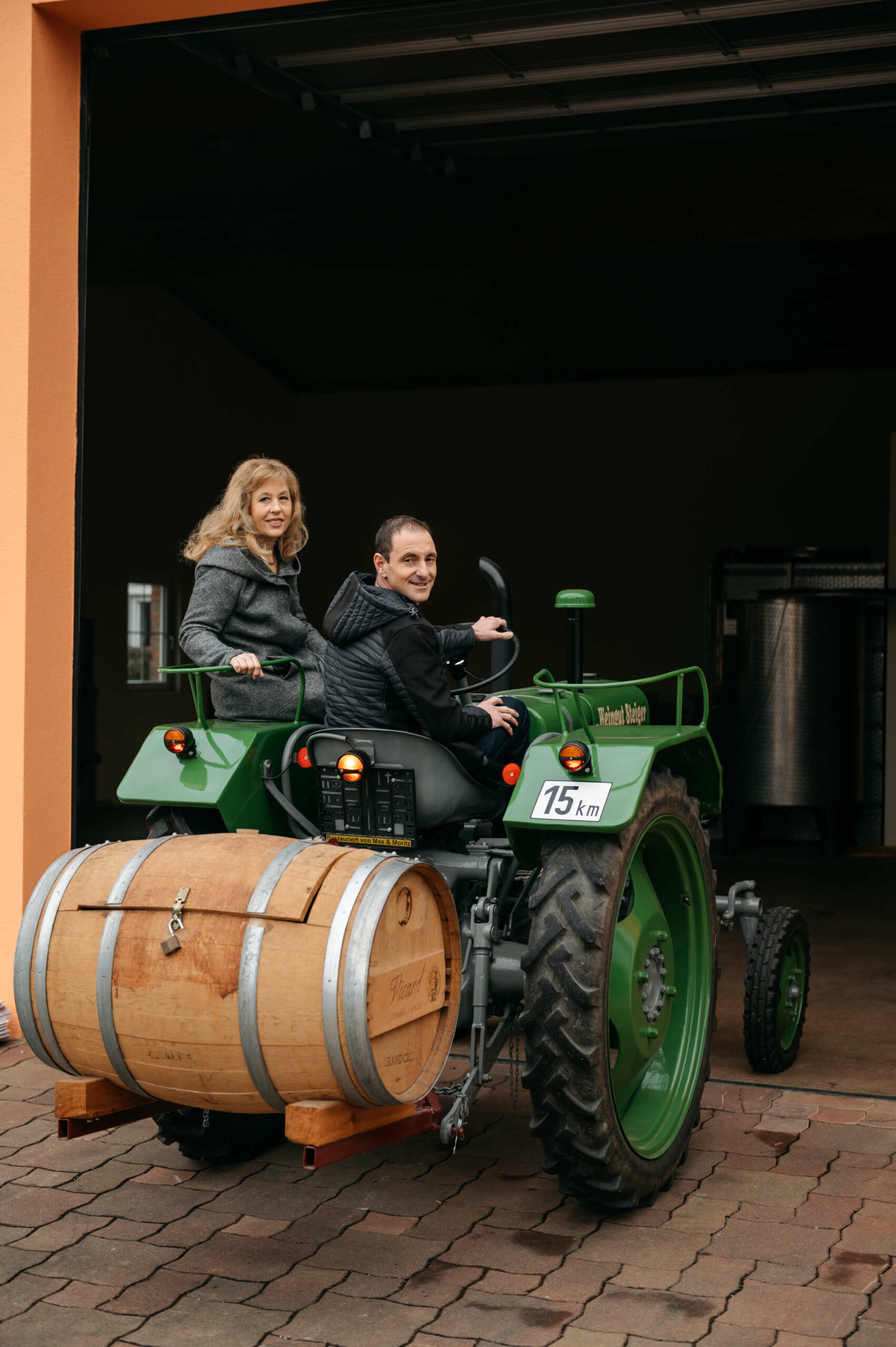 Herr und Frau Steiger sitzen auf einem grünen Traktor mit eine Weinfass